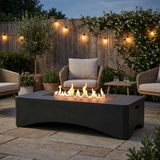 A NatureScape Living 56" Rectangular Gas Fire Pit Table with flames illuminates an inviting patio with outdoor seating at dusk.