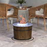 A NatureScape Living Smokeless Fire Pit with a wood barrel base and metallic top burns brightly on a patio, flanked by wicker chairs.