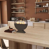 A black round bioethanol fire pit with dancing flames sits on a light wood table in a warm-toned modern kitchen.