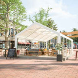 A white NatureScape Living Heavy Duty Party Tent 16x32FT shelters outdoor patio furniture on a raised platform in a town square.