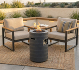 NatureScape Living's 26" Outdoor Gas Patio Fire Pit, dark grey with wavy texture, glows on a rug between two patio chairs.