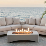 A modern minimalist rectangular propane fire pit table with glowing flames, surrounded by outdoor sofas on a patio by the ocean.