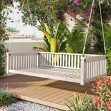 A white TOPMAX wood porch swing bed with slats hangs by ropes on a deck, surrounded by lush purple-flowering plants.