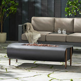 The VERNON Rectangle Fire Pit, dark gray with lava rocks and wooden legs, sits on a stone patio with an outdoor sofa.